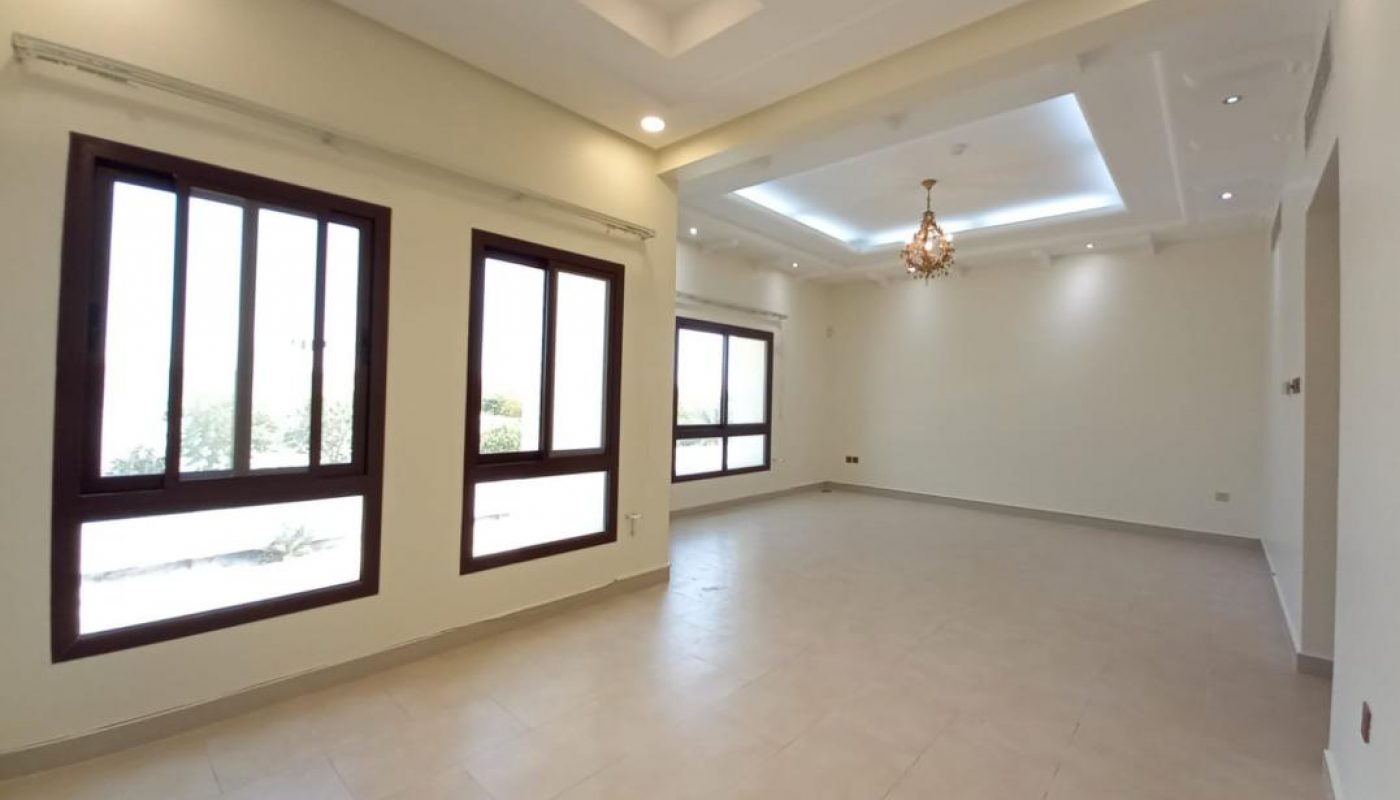Spacious, empty room with large windows, beige tiled floor, cream walls, recessed ceiling lights, and a chandelier. Natural light fills the space.