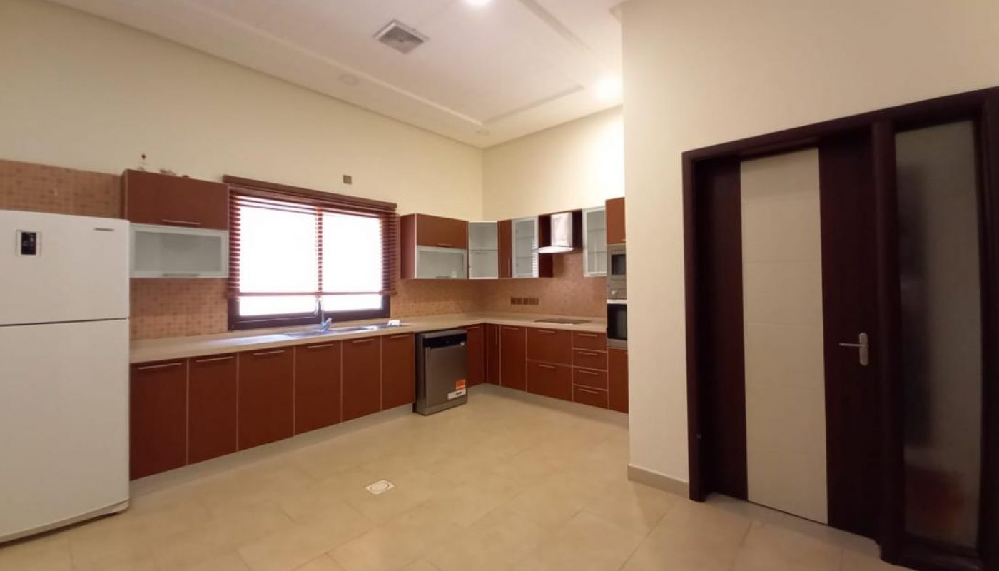 Modern kitchen with brown cabinets, white countertops, a refrigerator, dishwasher, oven, large window, and beige tile flooring. Sliding door is visible on the right side.
