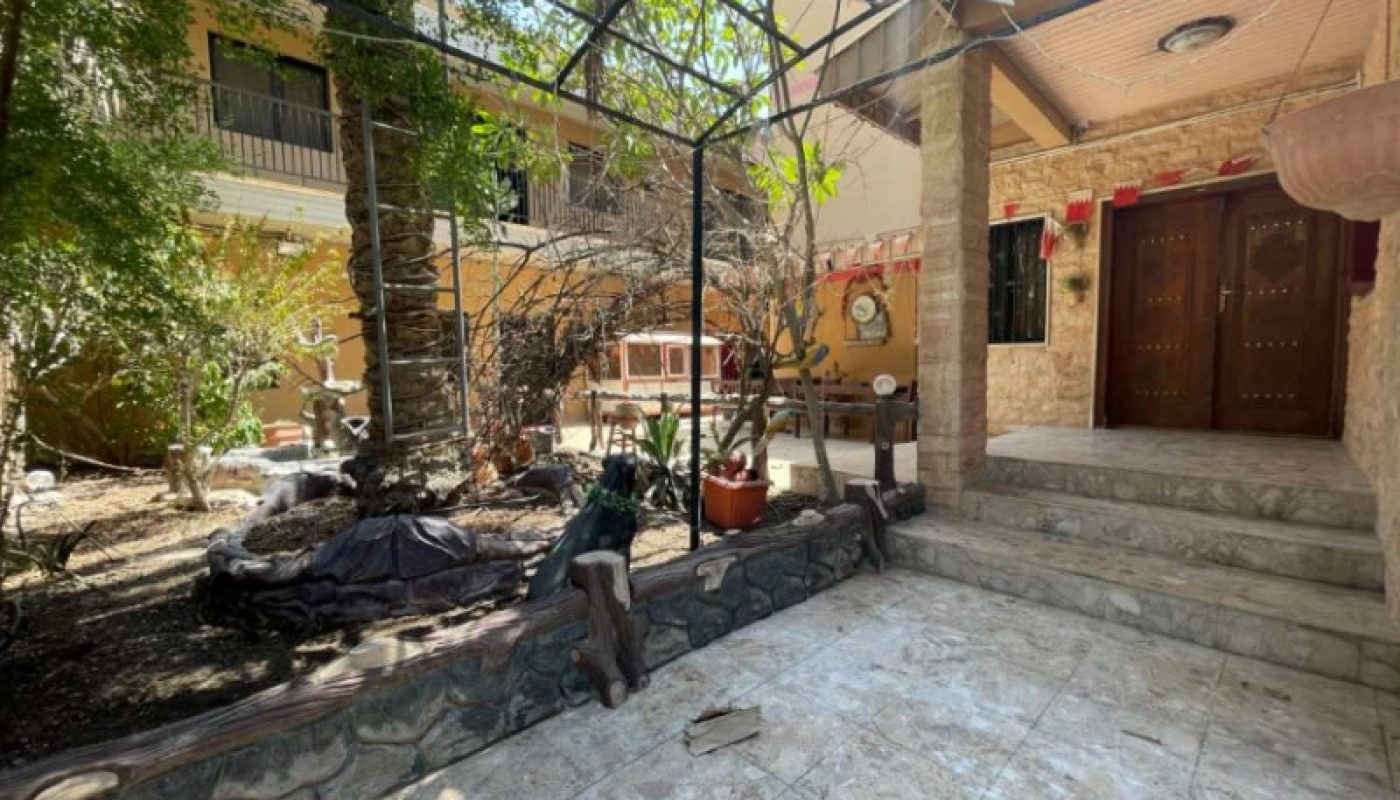 A stone-tiled courtyard with potted plants, a tree, and a small garden area next to a house entrance with double wooden doors and stone steps.