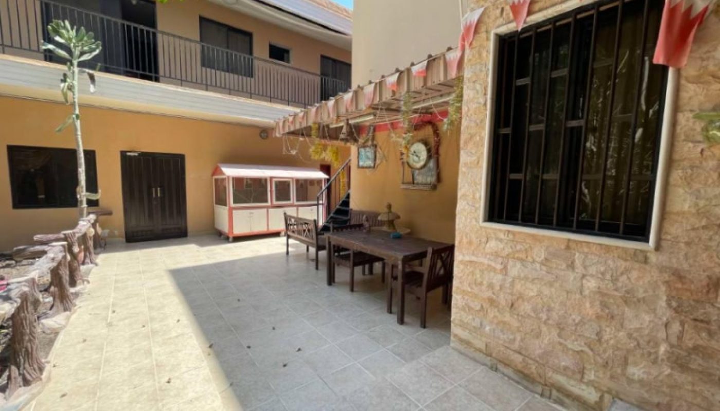 A tiled courtyard with a table and chairs, wall clock, window with bars, string of triangular flags, and a small playhouse near a building with two floors.