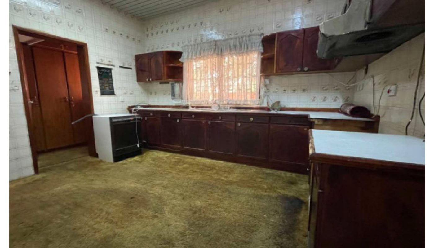 A spacious kitchen with brown carpet, dark wood cabinets, white tiled walls, a stove, and a large window with lace curtains.