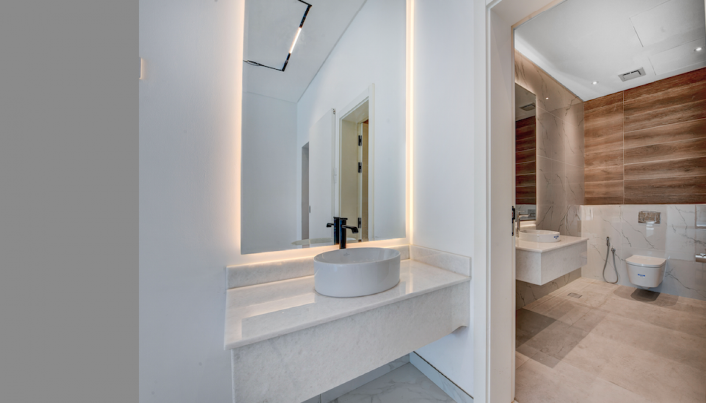 Modern bathroom with a round vessel sink, large illuminated mirror, and adjacent room featuring a wall-mounted toilet and wood accent wall.