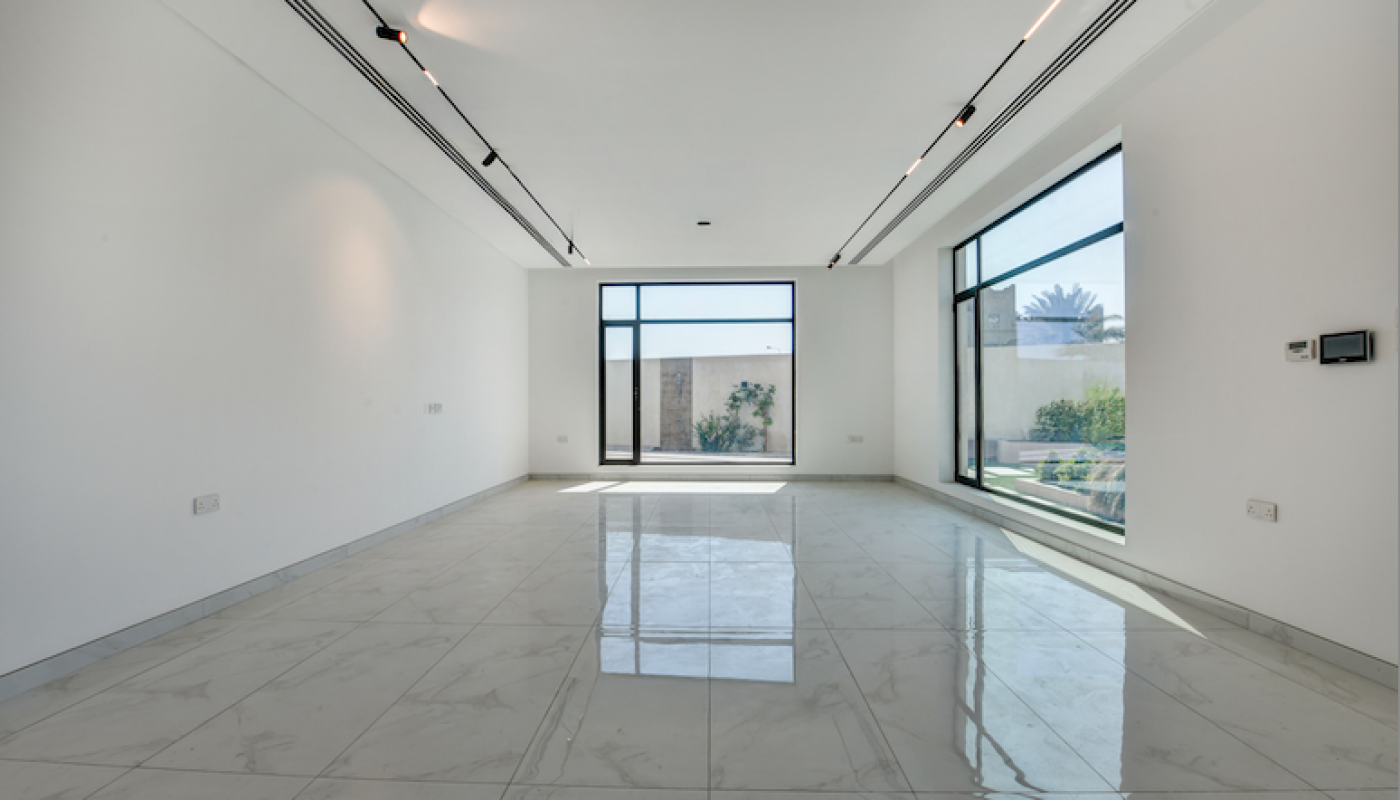 Spacious empty room with white marble floors, large windows, and white walls. Sunlight enters through the windows, illuminating the modern, minimalist interior.