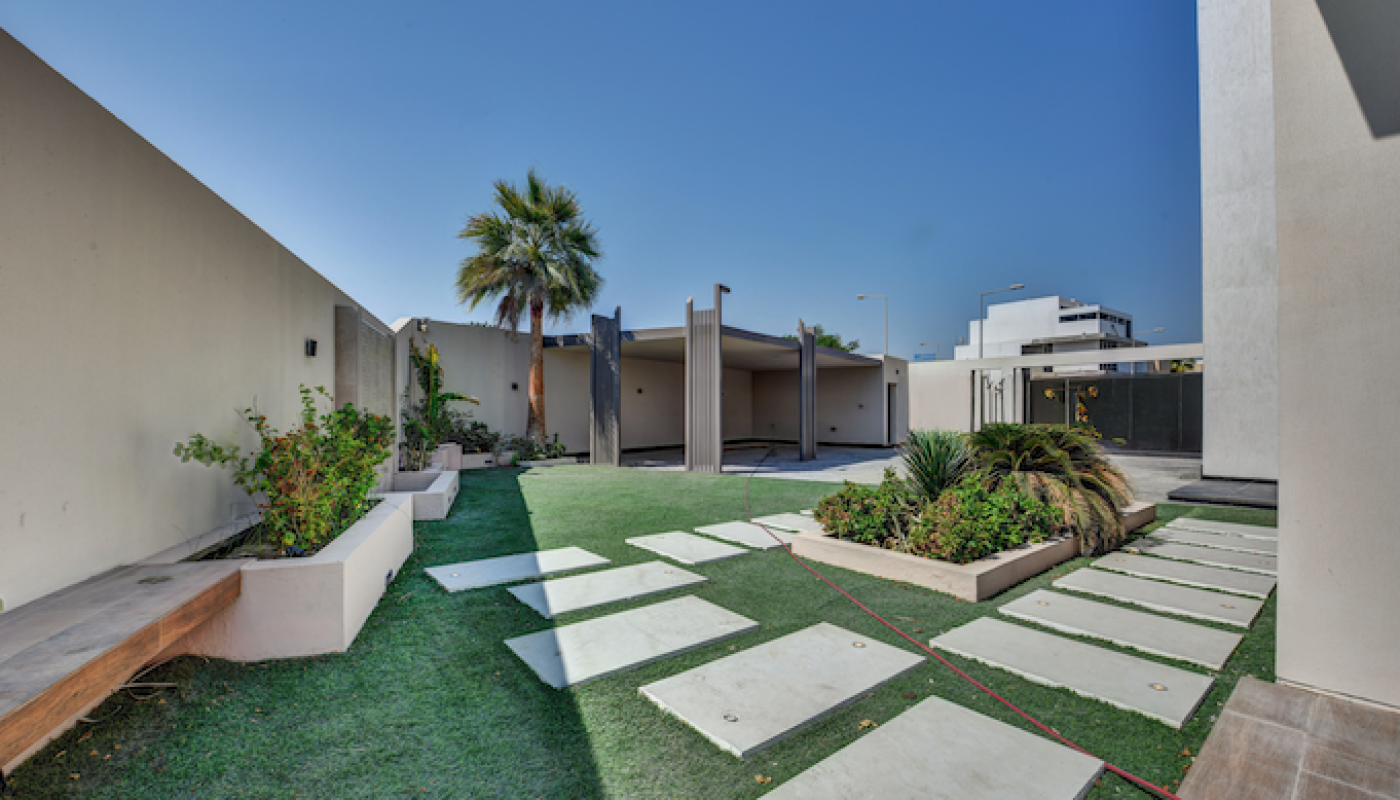 Modern outdoor courtyard with green lawn, stepping stone pathway, raised garden beds, a palm tree, and a covered seating area, surrounded by high white walls under a clear blue sky.