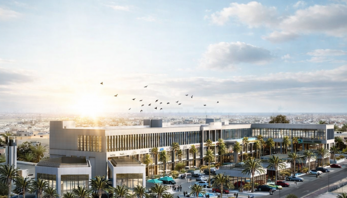 A modern commercial complex with large glass buildings, palm trees, parked cars, and people walking, set along a curved road under a partly cloudy sky at sunrise.