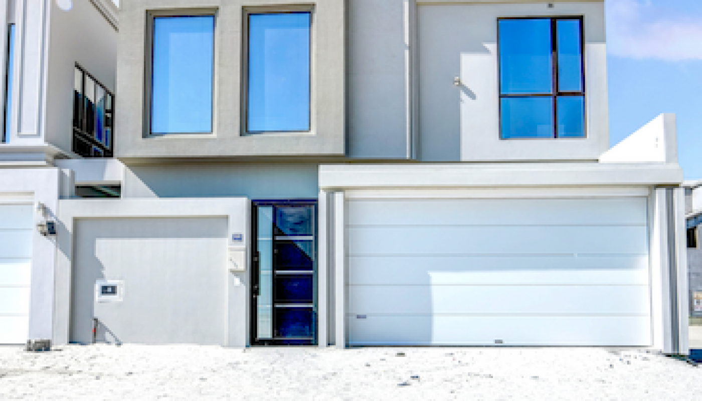 Modern two-story house with large windows, white exterior, and two garage doors, set against a bright blue sky with minimal landscaping in front.