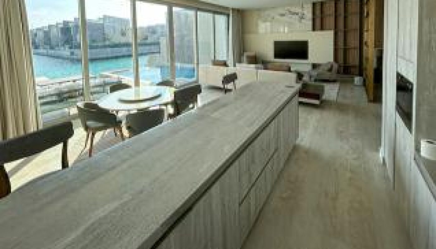 Modern open-plan living space with a long kitchen island, dining table, large windows, and a seating area overlooking a waterfront city view.