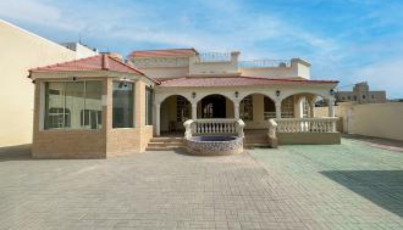 Single-story house with a large paved courtyard, covered porch, arched columns, and a detached glass-walled room on the left under a blue sky.
