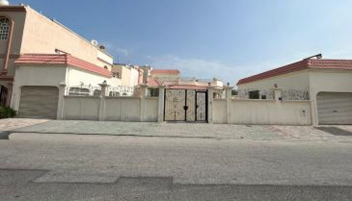 A wide, gated residential house with a beige exterior, red-tiled roof, and large garage doors, viewed from across a paved street under a clear blue sky.