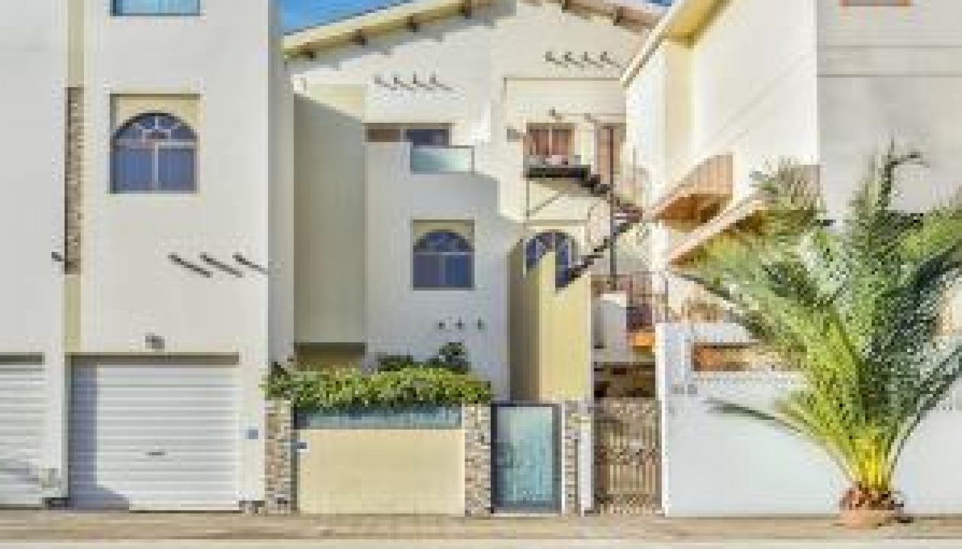 Three modern, light-colored houses with large windows and balconies stand side by side along a street under a blue sky.