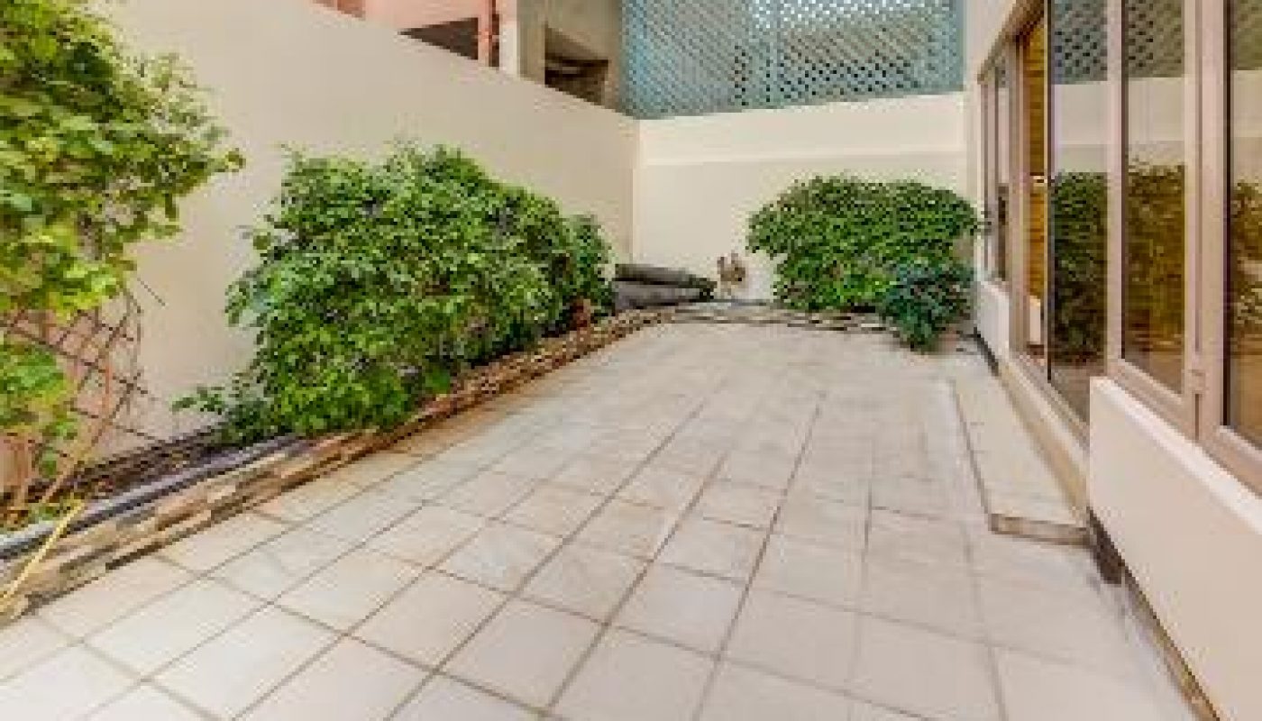 Rectangular tiled patio bordered by green shrubs, with light-colored walls on both sides and glass doors on the right. Apartments and an air conditioning unit are visible above.