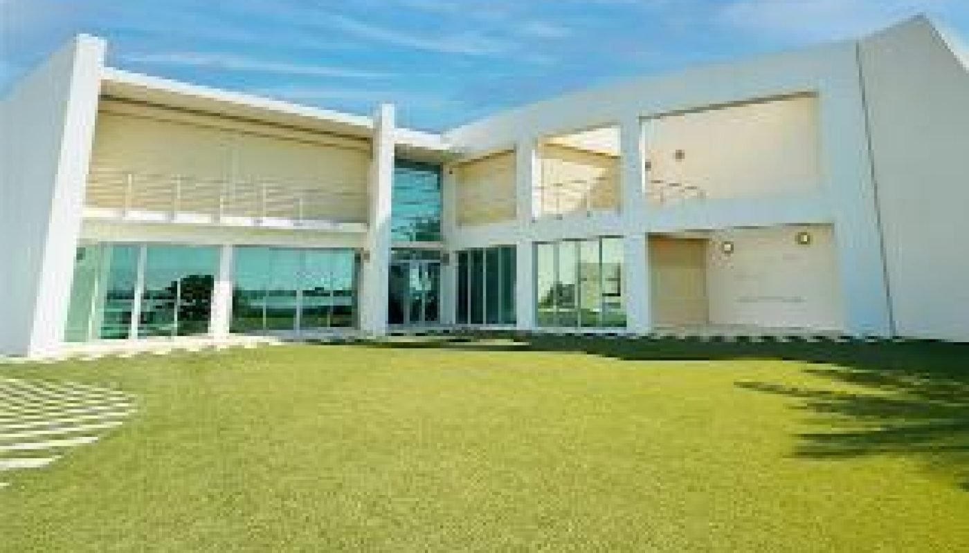 Modern white building with large windows and balconies, situated on a neatly manicured lawn under a blue sky with scattered clouds.