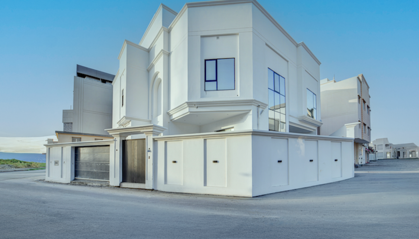 A modern two-story white house with large windows and a flat roof stands on a wide, empty street under a clear blue sky.