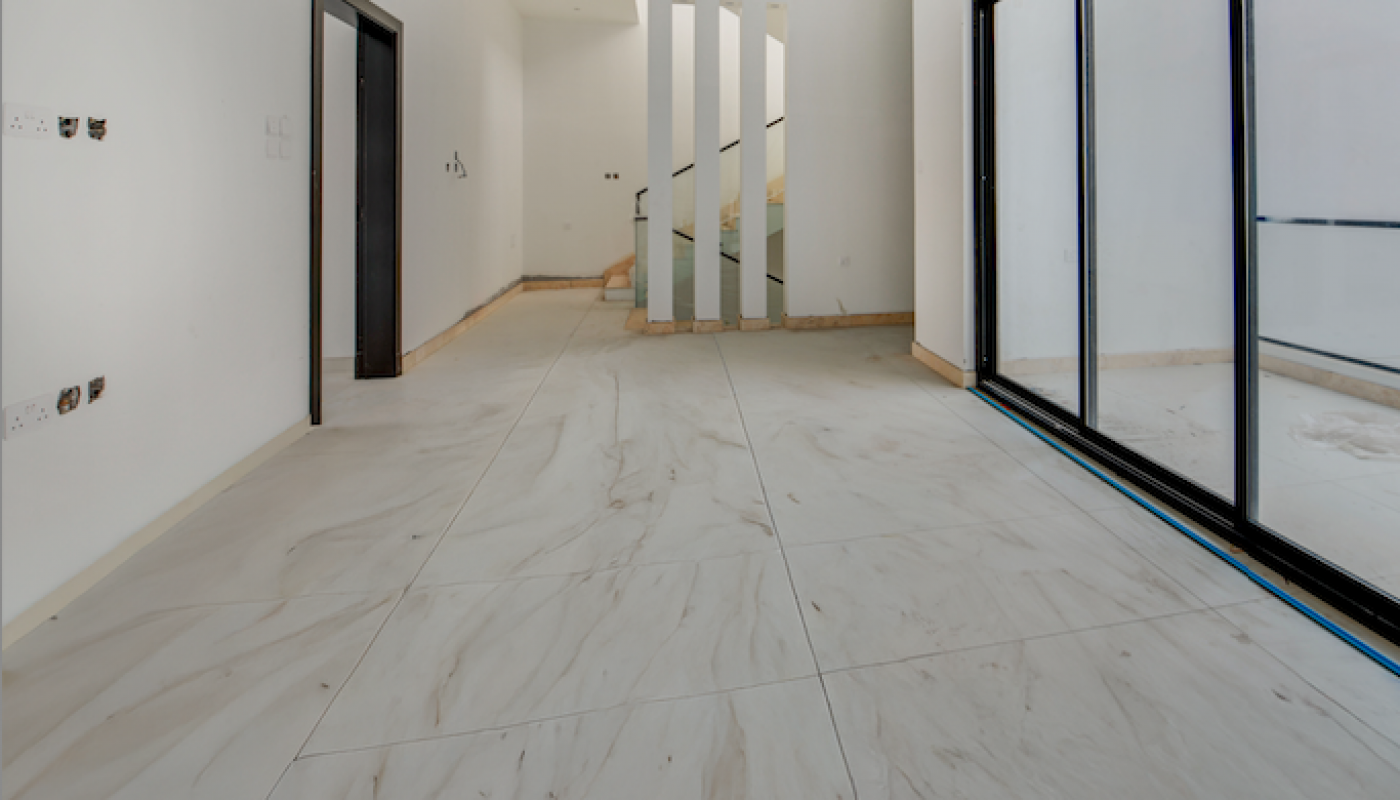 Spacious, unfurnished modern interior with large white tiles, glass sliding doors on the right, a dark door on the left, and staircase with vertical balusters in the background.