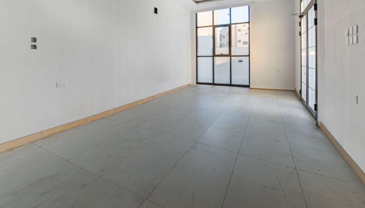 Empty, unfurnished room with large floor tiles, white walls, recessed ceiling lights, and floor-to-ceiling windows letting in natural daylight.