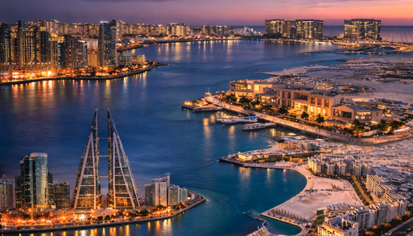 Aerial view of a coastal city at dusk, with illuminated modern buildings, waterfront promenades, and calm blue water reflecting city lights.