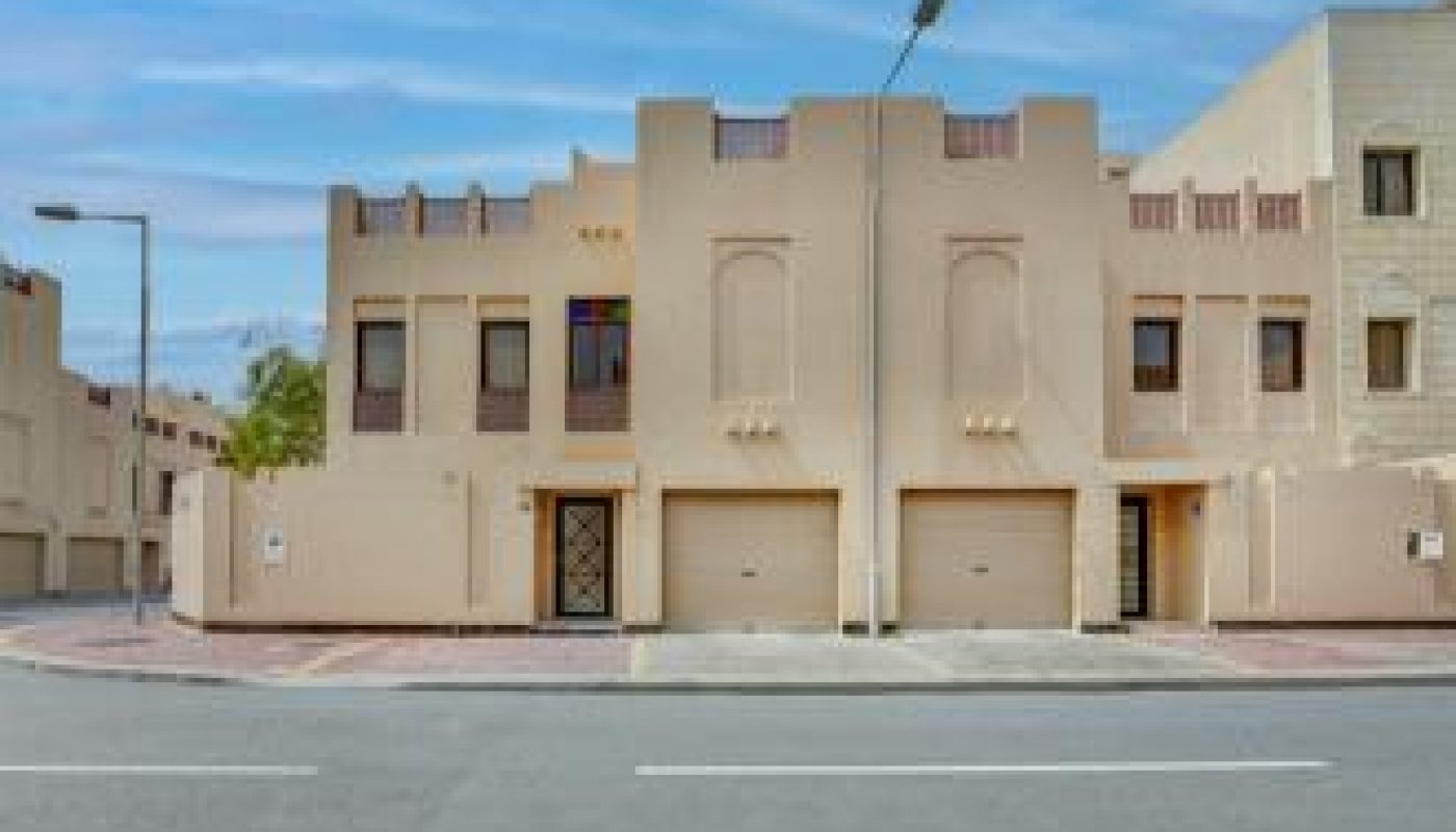 Two beige, modern townhouses with flat roofs and minimal architectural details face a wide, empty street under a blue sky with scattered clouds.