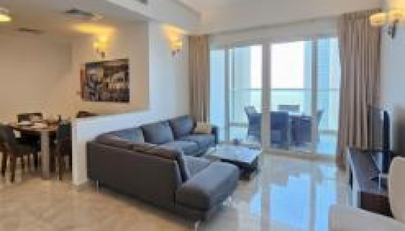 Modern living room with gray sectional sofa, armchair, TV, dining area, large windows with curtains, and a balcony visible outside. Marble tile floor and contemporary ceiling lights.