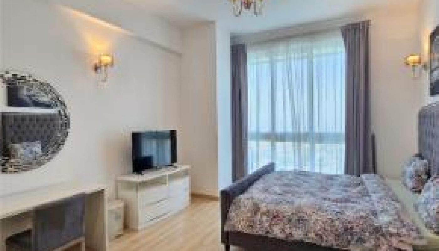 Modern bedroom with a large bed, floral bedding, a TV on a white dresser, a vanity with a round mirror, wood flooring, and large window with sheer curtains.