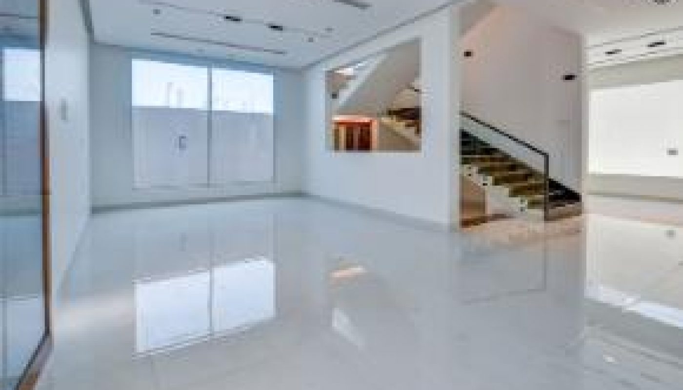 Spacious, empty room with white floors and walls, large windows, and a modern staircase with glass railings leading to an upper level.
