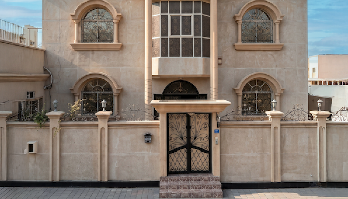 Two-story beige house with arched windows, a central balcony, and a gated entrance set against a clear blue sky.