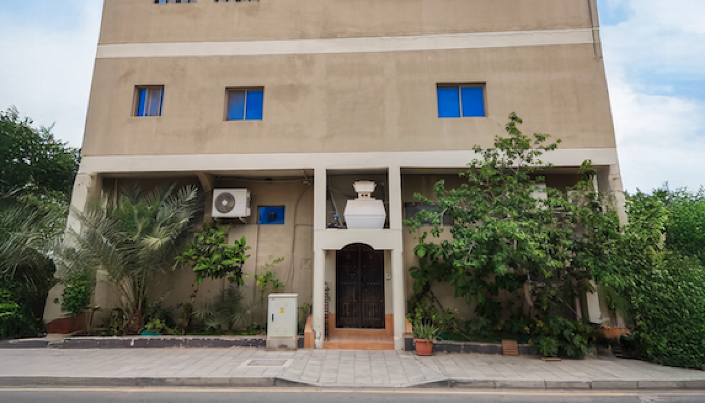 Three-story beige building with arched windows, blue accents, rooftop enclosure, and greenery along the entrance, situated next to a paved road.