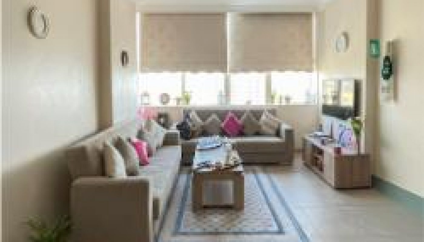 A spacious living room with beige walls, two sofas, a coffee table, a TV unit, and large windows with roller blinds letting in natural light.