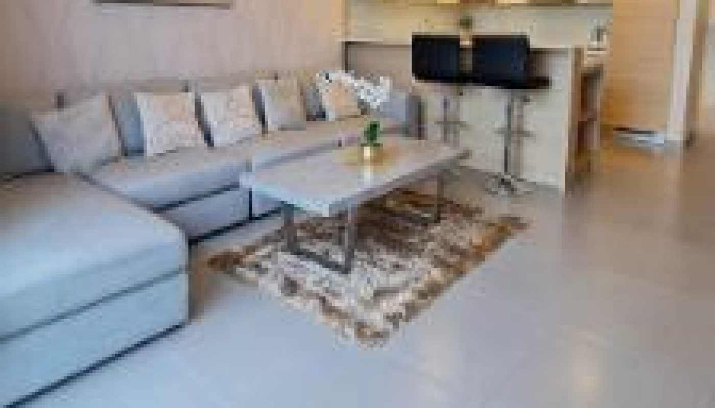 Modern living room with a light gray sectional sofa, coffee table, small rug, wall mirror, TV, and open kitchen area with bar stools in the background.