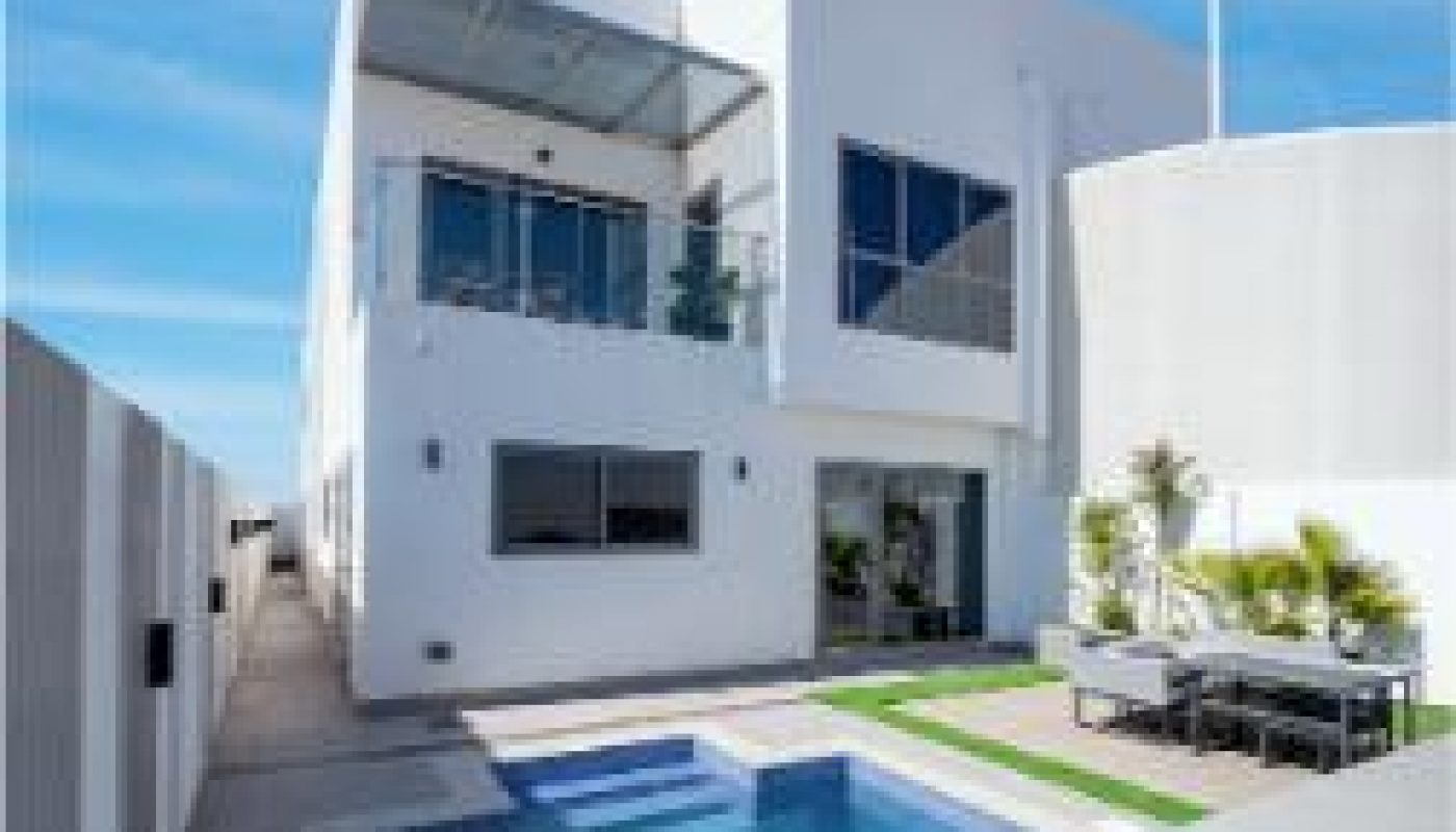 Modern two-story white house with large windows, a balcony, and a rectangular outdoor swimming pool beside a small patio dining area.