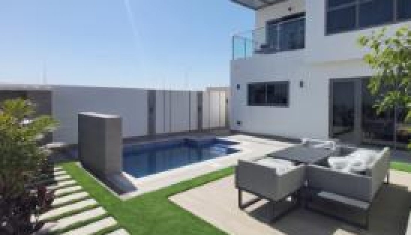 Modern outdoor patio with a small pool, lounge seating, green artificial grass, stone path, white pebbles, and a white two-story building under a clear blue sky.