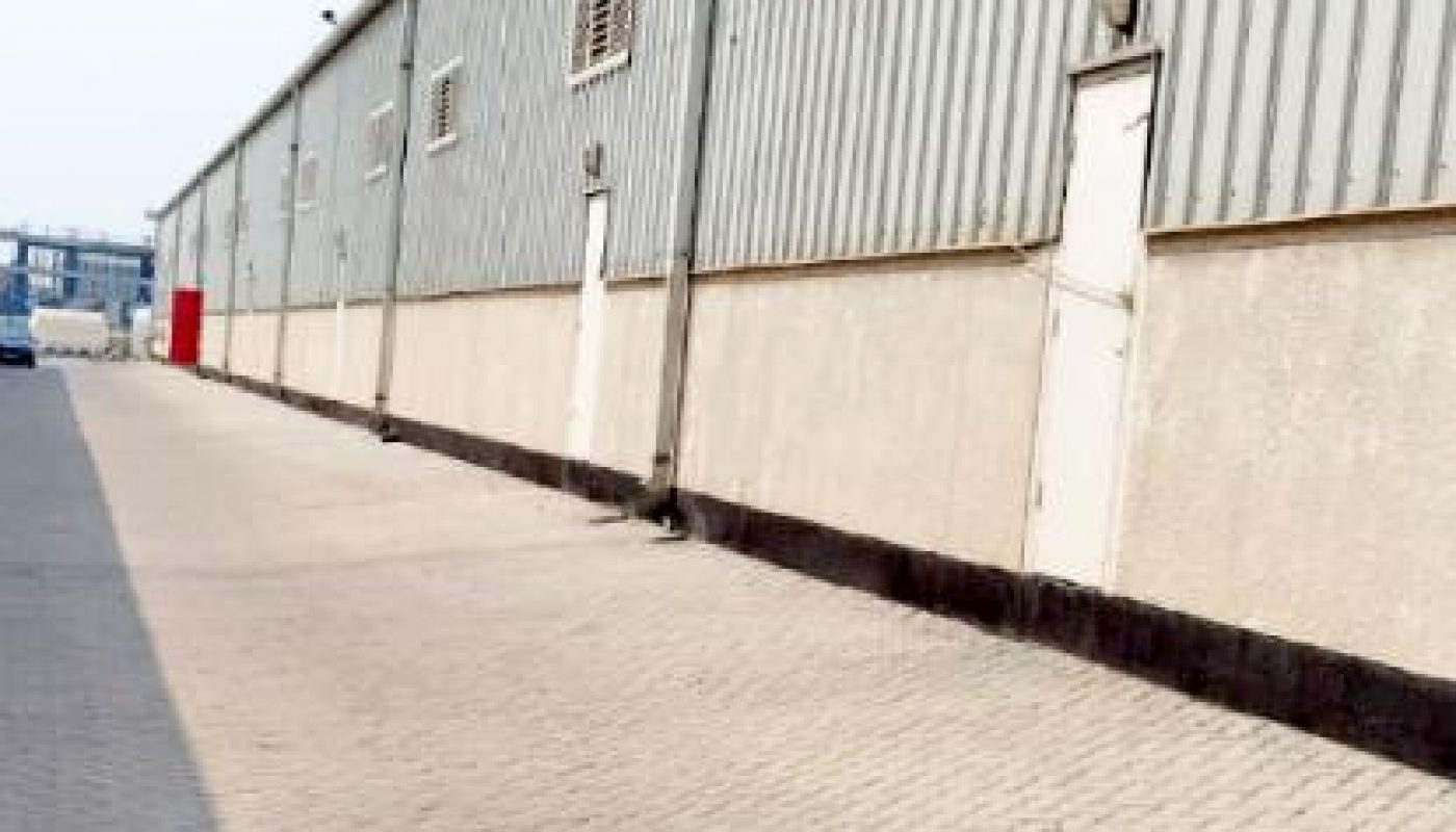 Long, light-grey industrial warehouse building with several white doors and ventilation units, situated on a paved brick driveway under a clear sky.