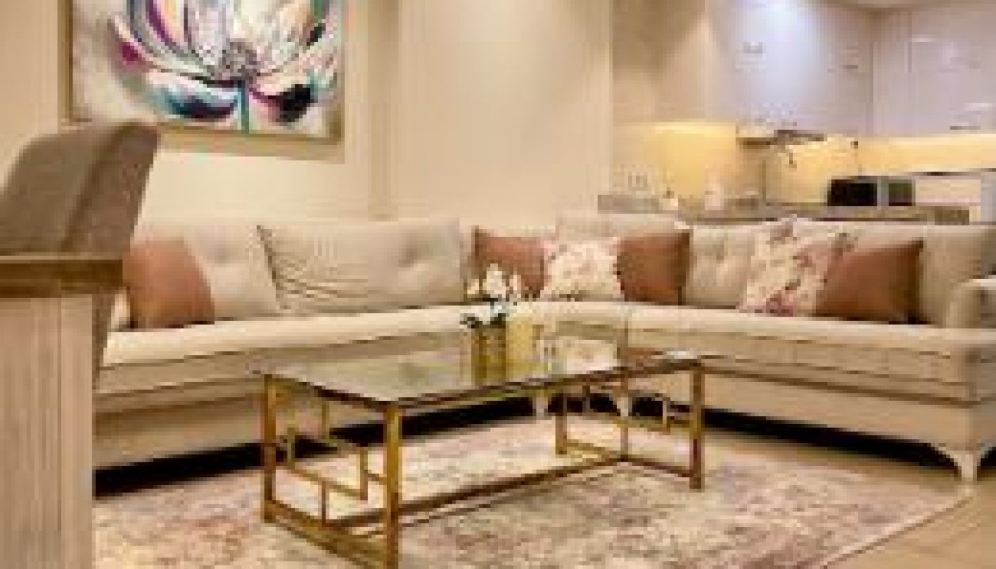 Modern living room with beige sectional sofa, glass coffee table, floral artwork, decorative pillows, and adjoining kitchen in the background. Neutral color scheme and warm lighting.