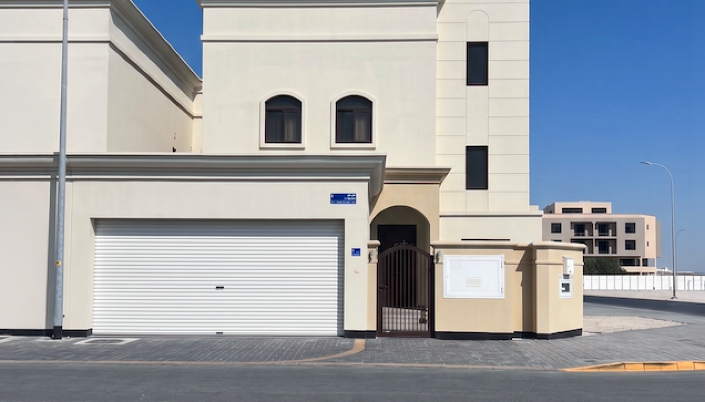 A modern two-story beige house with arched windows, a white garage door, and a small gate, situated on a paved street corner under a clear blue sky.