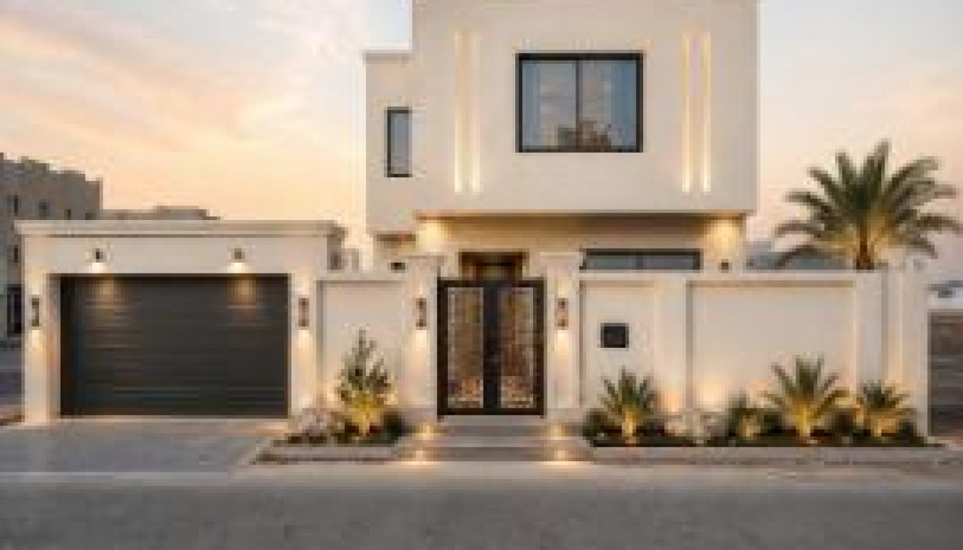 Modern two-story house with a flat roof, large black garage door, and decorative gate, illuminated by exterior lights at sunset, with a palm tree and plants in front.
