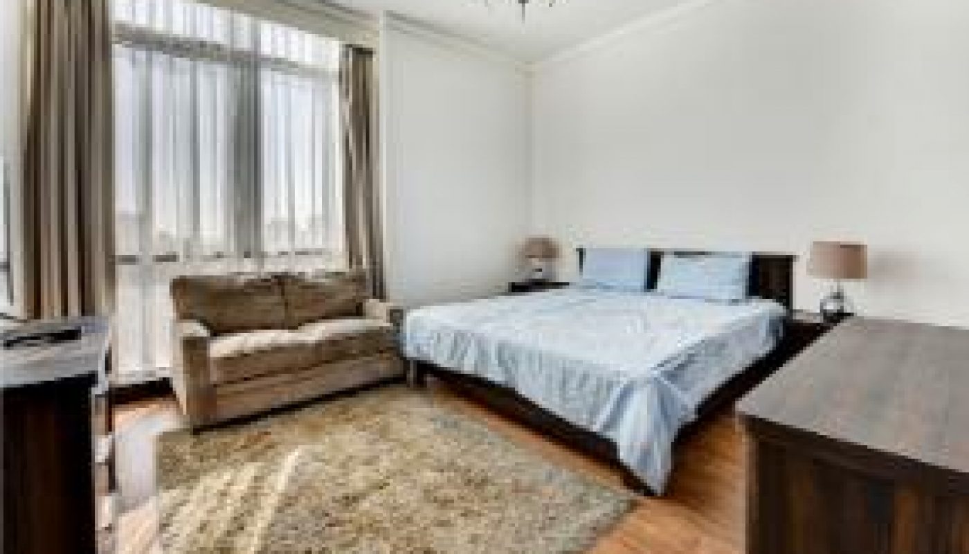 A bedroom with a double bed, brown sofa, wooden furniture, a rug, large window with curtains, and a ceiling light fixture.