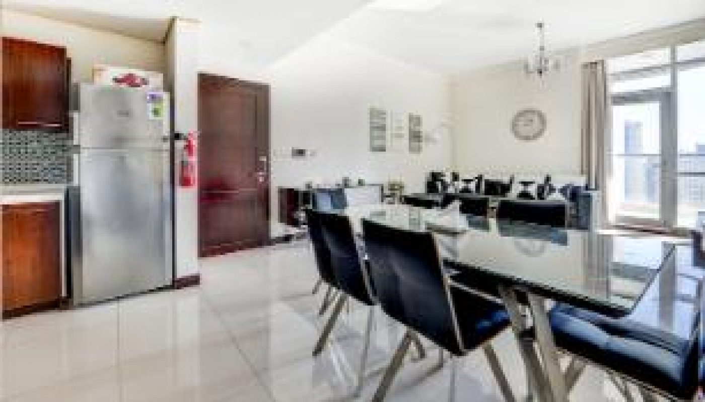 Modern dining area with black chairs, glass-top table, open kitchen, stainless steel fridge, and living room with sofa in a bright, spacious apartment.