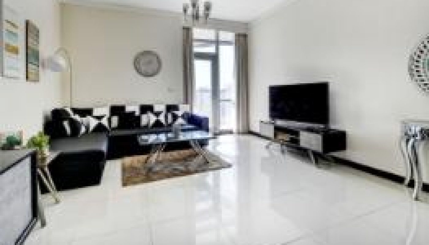 Modern living room with white tile floor, black sofas, glass coffee table, TV on a stand, wall art, and a chandelier. Large window lets in natural light.
