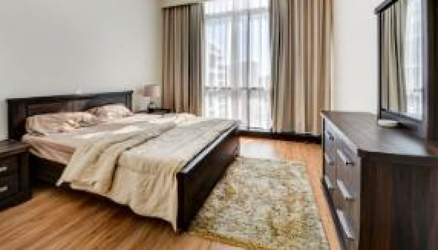 Modern bedroom with an unmade bed, wooden floor, area rug, dresser with mirror, nightstand, and large curtained window letting in natural light.