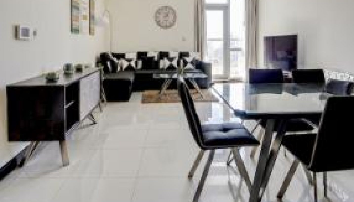 Modern living and dining area with a black sofa, glass dining table with four chairs, TV, console tables, wall clock, and artwork on white marble flooring.