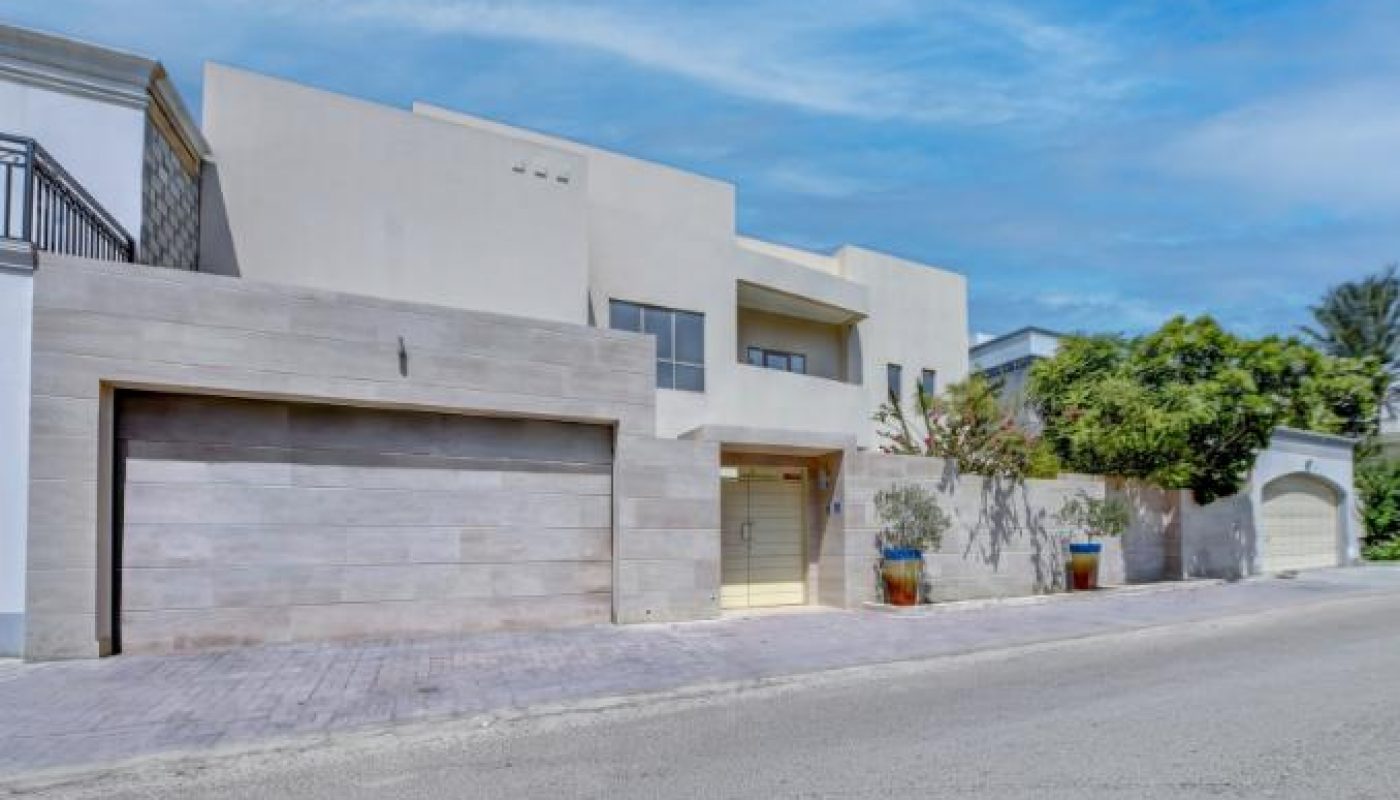 Modern two-story house with a flat roof, large garage doors, and minimalistic design, situated on a quiet street under a blue sky.