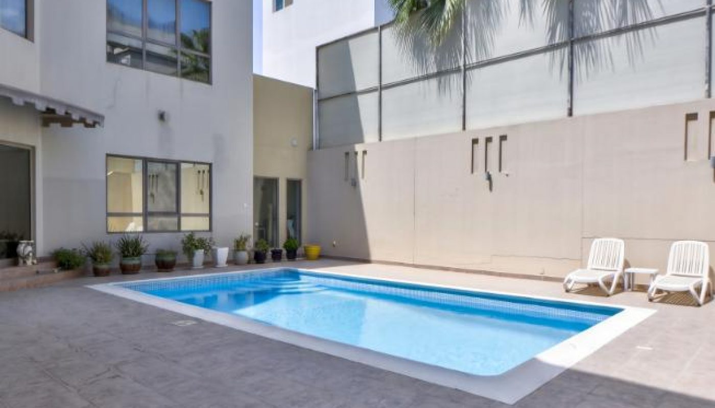 Rectangular outdoor swimming pool in a modern apartment courtyard, with two white lounge chairs, potted plants, and tall palm trees in the background.
