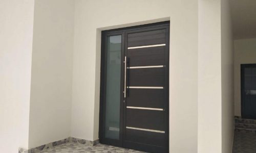 A modern black door with horizontal silver accents and a sleek vertical handle, set in a white wall, with a tiled entry area, perfectly complements the luxurious villa for sale in Maqaba. Another open doorway is visible to the right.