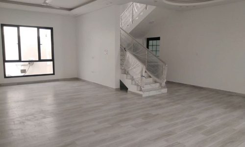 A spacious empty room in a villa for sale in Maqaba, featuring light gray wooden flooring, white walls, and a staircase leading to an upper level. There is a large window on one side and recessed ceiling lights.