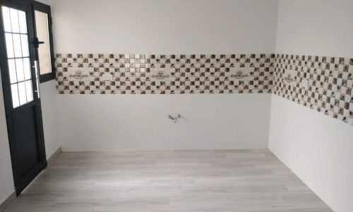 A room in a charming villa for sale in the Maqaba area, featuring white walls, a black-framed window, and a checkered tiled backsplash. The light wood floor and exposed water pipes suggest it may be a kitchen area, blending functionality with modern aesthetics.