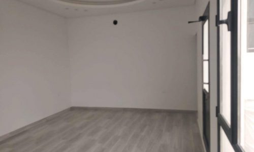 An empty room with white walls, a gray floor, recessed lighting, a circular ceiling feature, and two large windows on the right, located in an elegant villa for sale in Maqaba.