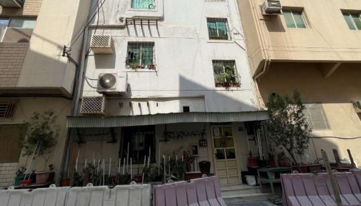 A four-story residential building with barred windows and plants outside, partially blocked by construction barriers on a sunlit street.