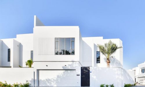 A modern white two-story luxury villa with large windows, a garage, and a black gate. There's a tall palm tree adjacent to the 650 SQM house, and the sky is clear and blue.