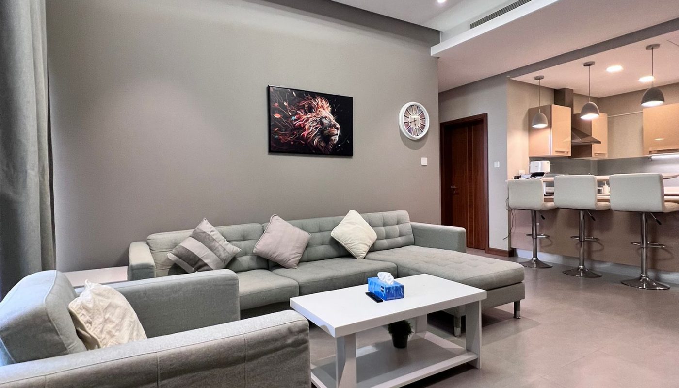 Modern living room with grey sofas, a white coffee table, wall art, and an open kitchen area with bar stools in the background. The space is well-lit and minimalistic.