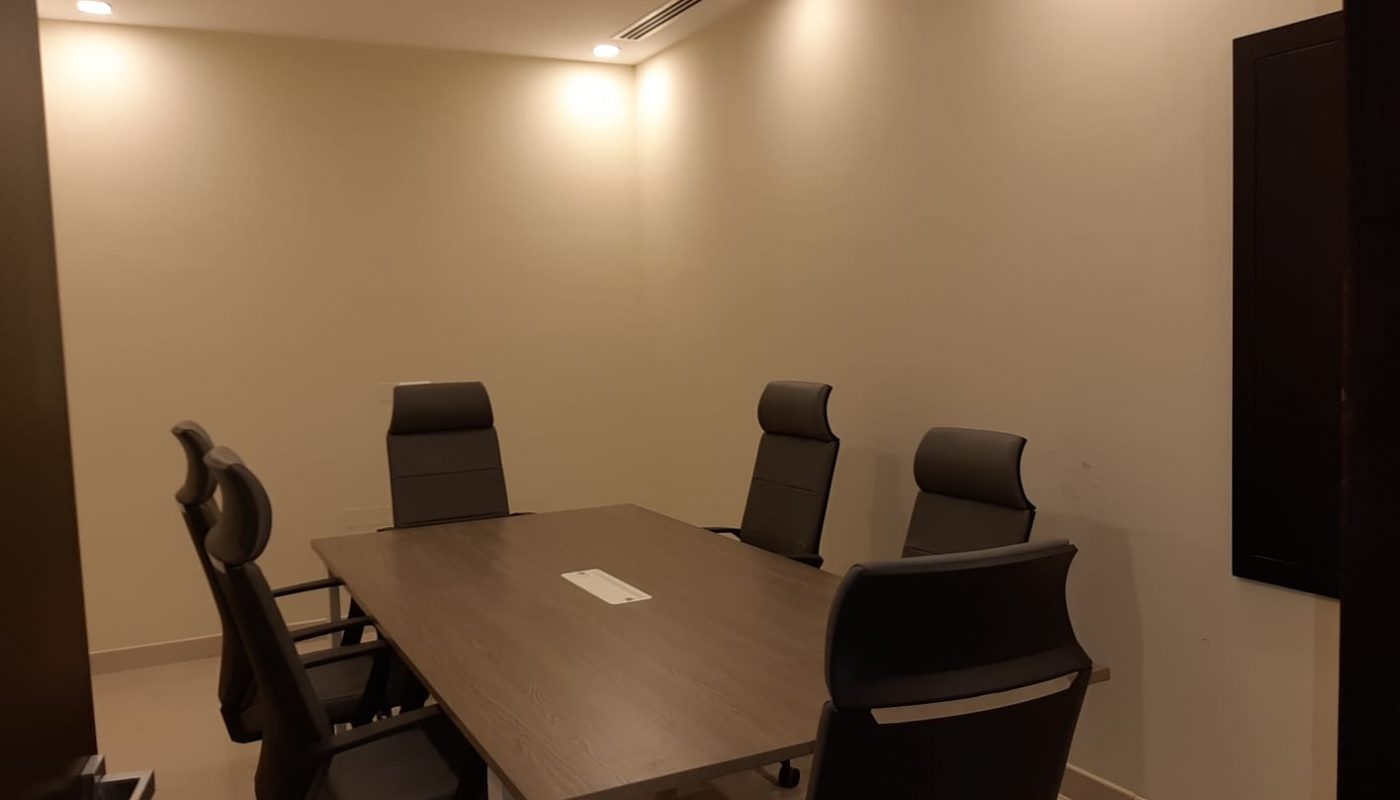 A small conference room with a rectangular table and five black office chairs arranged around it, lit by ceiling lights.