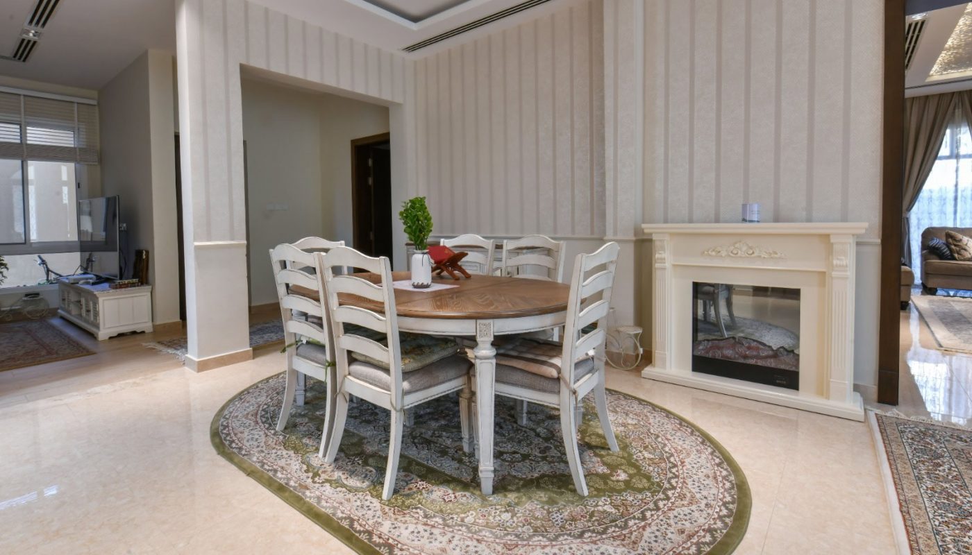 Dining room with a round wooden table, six white chairs, and a decorative rug. A fireplace is on the right, and adjacent rooms are visible through open doorways.